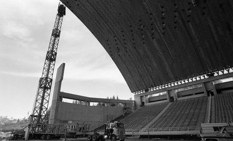 Black and white photograph of an arched roof under construction, attached to concrete stands.