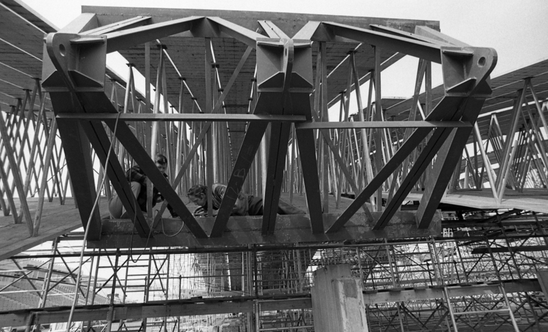 Black and white photograph of a man working inside two arched wooden beams separated by metal trusses.