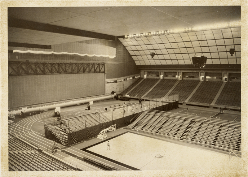 Black and white photograph of the interior of an enclosed stadium, with additional stands set up on the field surrounding a basketball court.