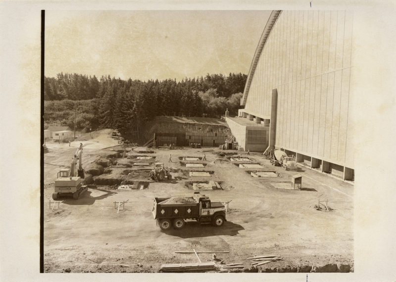 Black and white photograph of a truck and other heavy machinery on a construction site next to a building with an arched roof.