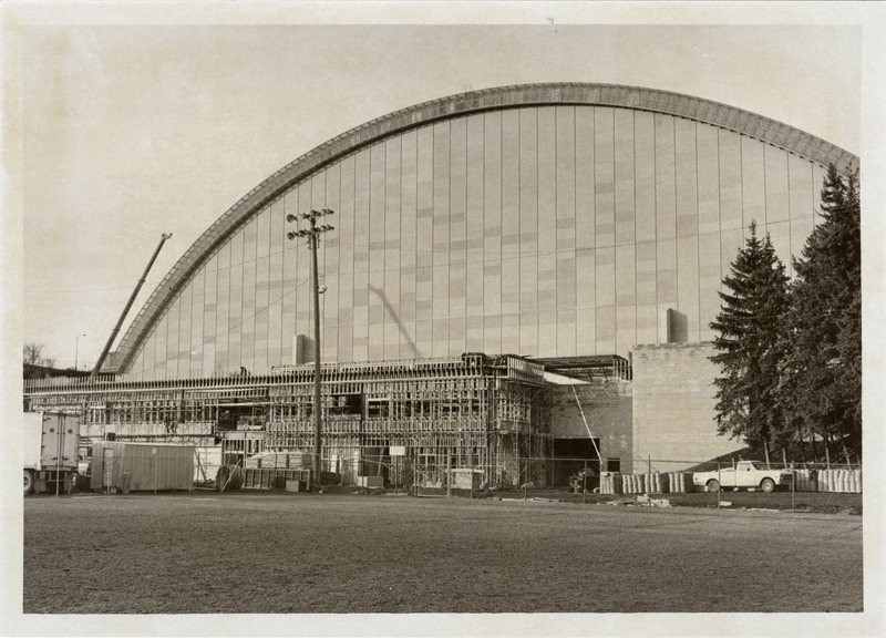 Black and white photograph of a low rectangular building under construction next to a building with an arched roof.
