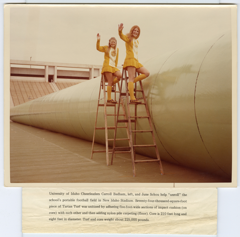 Color photograph of two women in yellow and white cheerleader uniforms standing on ladders next to a long, green roll of flooring. Empty stadium stands are in the background. Text below the photograph reads: "University of Idaho Cheerleaders Carroll Badham, left, and June Schou help "unroll" the school's portable football field in New Idaho Stadium. Seventy-four-thousand-square-foot piece of Tartan Turf was unitized by adhering five-foot-wide sections of impact cushion (on core) with each other and then adding nylon pile carpeting (floor). Core is 210 feet long and eight feet in diameter. Turf and core weigh about 225,000 pounds."