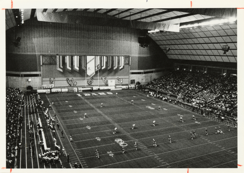 Black and white photograph of a football game taking place in an enclosed stadium with an arched roof. People sit in the stands on the left and right.