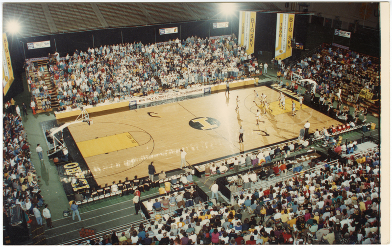 Color photograph of a well-illuminated basketball court with a game in progress. People fill the stands.