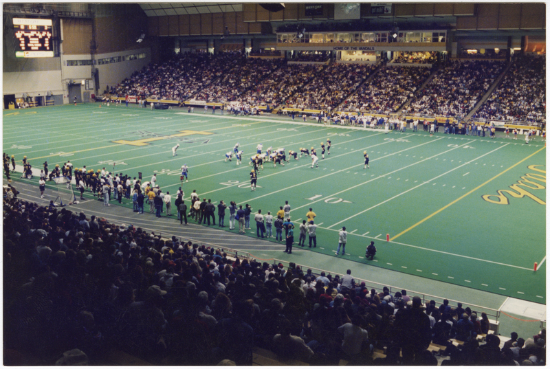 Color photograph of a football game in progress in an enclosed stadium. People sit in the stands in the foreground and background.