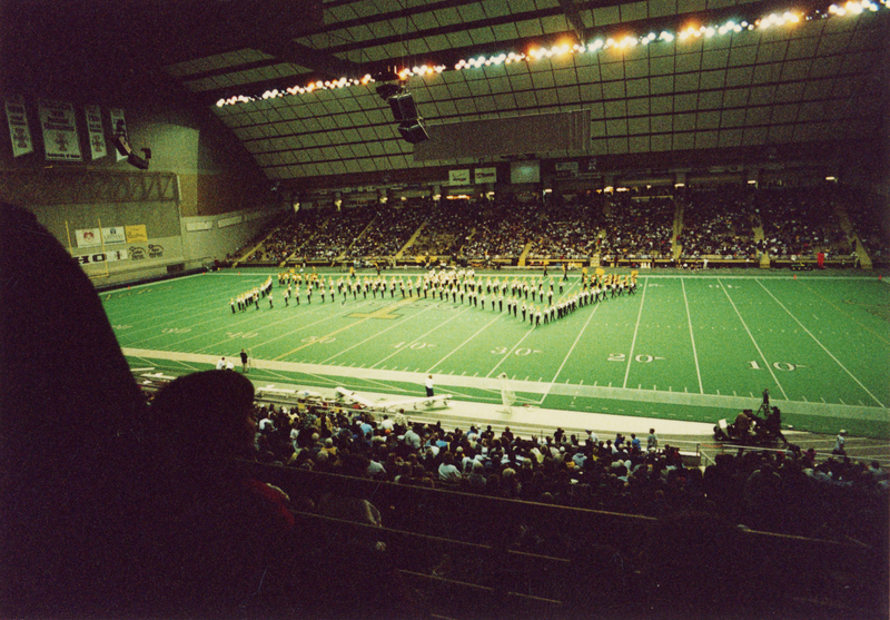Color photograph of a marching band in formation on an football field. People sit in the stands in the foreground and background.