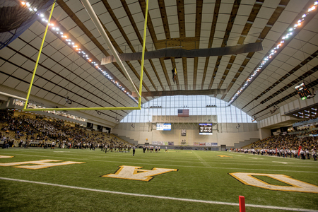 Color photograph of an enclosed football stadium. Football players stand on the field and the stands are partially occupied with spectators.
