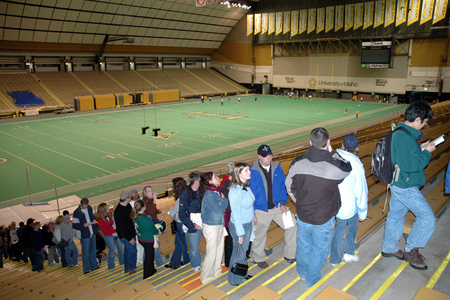 Color photograph of a line of people standing on the steps of an enclosed stadium. A few people are on the field. The stands are otherwise empty.