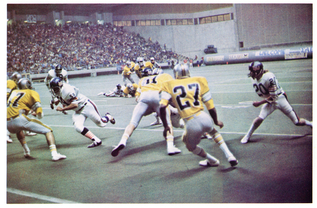 Color photograph of football players in two different styles of football uniforms running on a football field. In the background are stands crowded with people.