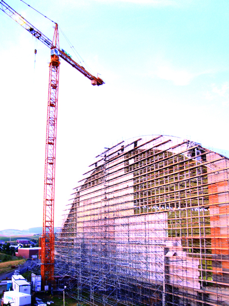 Color photograph of the end wall of a building covered in scaffolding. Next to it stands a construction crane and other construction equipment.