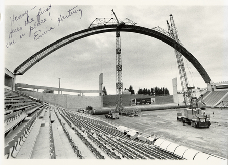 Black and white photograph of an open air stadium. Construction cranes maneuver a wooden arch to attach it to the upper wall of the stadium.