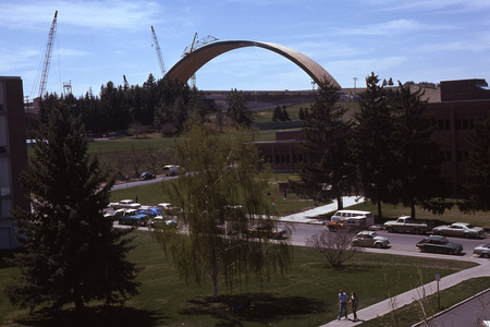 Color photograph of a grassy area surrounded by brick buildings. In the distance is a large arched roof under construction.