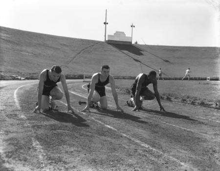 Black and white photograph of three men crouching on a running track. In the background is a grassy hillside.