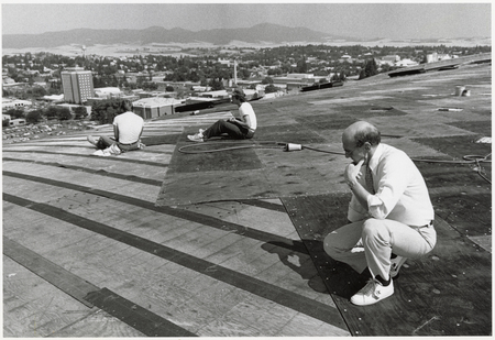 Black and white photograph of a man kneeling on the arched roof of a building. Two other people and some tools also sit on the roof. In the background are buildings and hills.