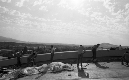 Black and white photograph of several people using tools to work on the top of an arched roof. In the background are buildings and hills.