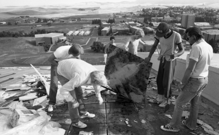 Black and white photograph of several people standing on the arched roof of a building. One holds up a stained section of wood. In the background are buildings and hills.