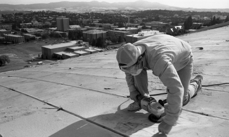 Black and white photograph of a person sanding the roof of an arched building. In the background are buildings and hills.