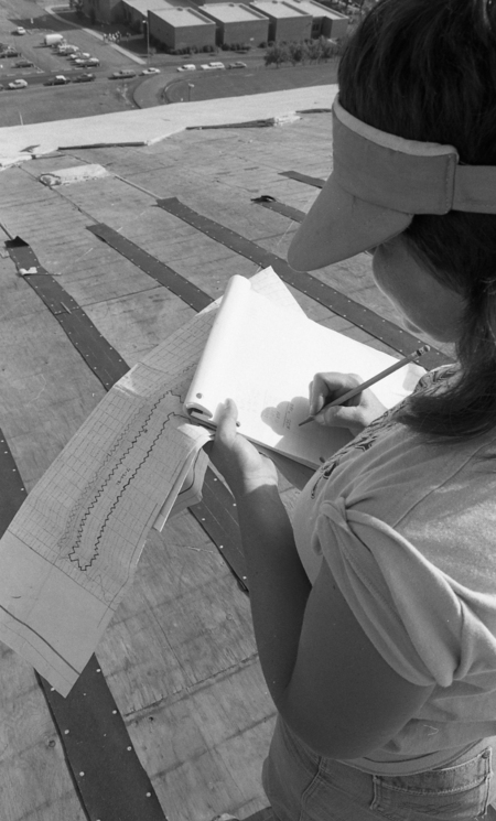 Black and white photograph of a woman writing on a pad of paper while standing on an arched wooden roof. She holds a piece of paper that appears to be a diagram of the roof.