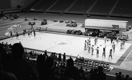 Black and white photograph of people standing on a basketball court. In the foreground are people sitting in stands. In the background is a truck and other materials sitting on the rest of the stadium field.
