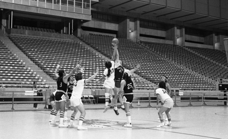 Black and white photograph of basketball players in two different uniforms, jumping for the basketball on a court in an enclosed stadium.