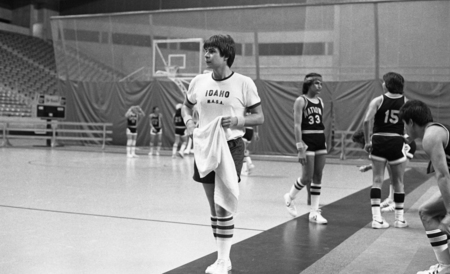 Black and white photograph of basketball players in two different uniforms walking on a basketball court. The white uniform shirt reads "IDAHO N.A.S.A." and the black uniform shirt reads "NATION".