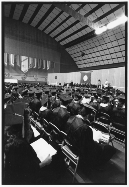 Black and white photograph of people wearing graduation robes and mortarboards sitting in rows inside an enclosed stadium with an arched roof.. In the background are more people sitting on a stage.