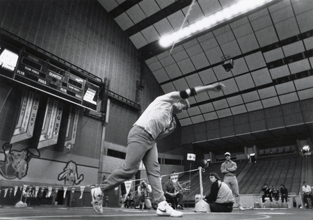 Black and white photograph of a javelin thrower standing on the field of an enclosed stadium. In the background are empty stands and several other people.