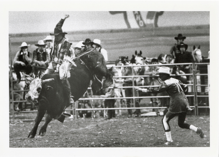 Black and white photograph of a person riding a bucking bull on a dirt field. Another person stands on the field nearby. In the background are spectators.