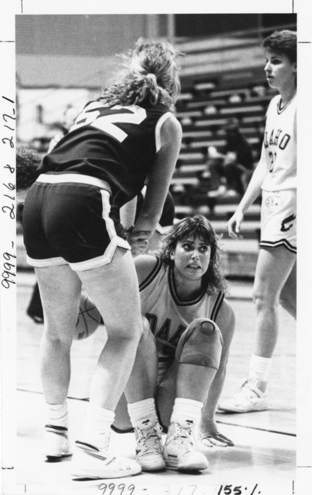 Black and white photograph of one basketball player helping another up off the floor. The women wear dark and light uniforms.