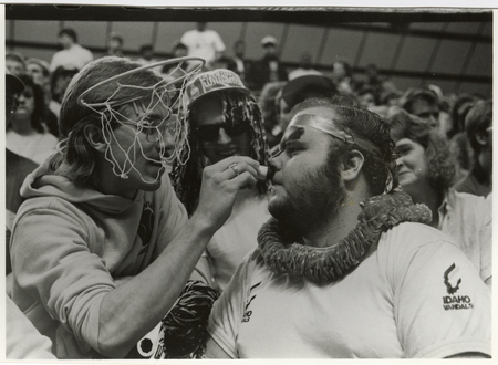 Black and white photograph of a person wearing a basketball hoop on his head and applying face paint to another person. In the background are spectators sitting in stands.
