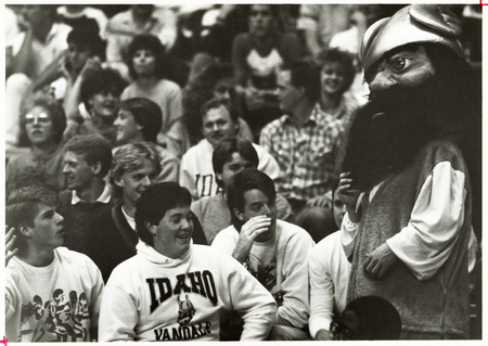 Black and white photograph of spectators sitting in stands interacting with a person in a mascot costume.