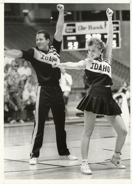 Black and white photograph of two people in cheerleader uniforms that read "IDAHO". In the background are spectators sitting in stands and a scoreboard.