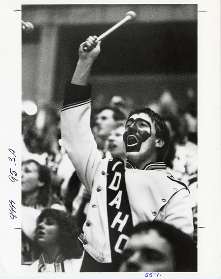 Black and white photograph of a person in a marching band uniform that reads "IDAHO." They hold a mallet and are surrounded by spectators in stands.