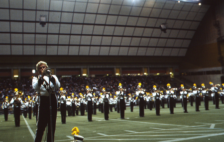 Color photograph of a trumpeter in a marching band uniform playing into a microphone. In the background are more marching band members and stands filled with people.