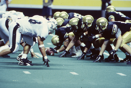 Color photograph of members of two football teams crouching and facing each other on a football field. One team wears white and black, the other wears gold and black.