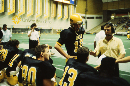 Color photograph of a man wearing a headset and talking to football players sitting and standing on the sidelines of a football field.