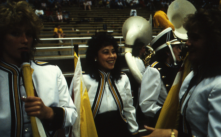 Color photograph of women wearing white, yellow, and black uniforms and holding white and yellow flags. In the background, people sit in stands.