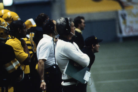 Color photograph of a man wearing a headset on the sidelines of a football field. Behind him are football players wearing black and yellow uniforms.