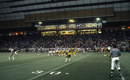 Color photograph of football players on an enclosed football field. In the foreground is a referee in a black and white uniform; in the background, spectators sit in stands.