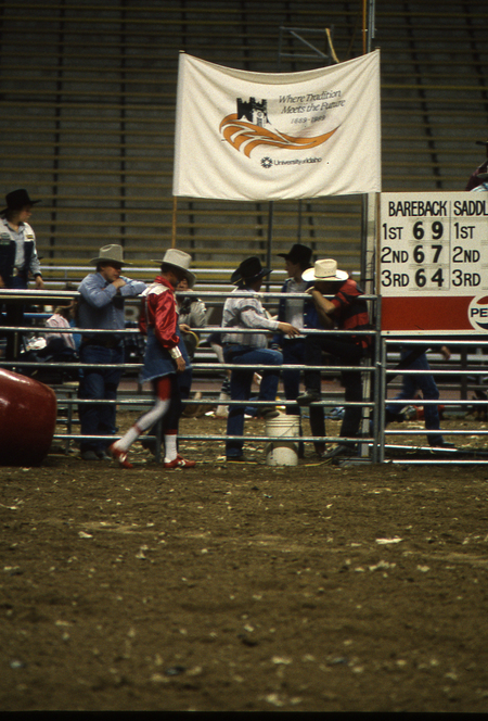 Color photograph of people standing along a metal fence on a dirt field inside of a stadium. A banner above them reads "Where Tradition Meets the Future University of Idaho".
