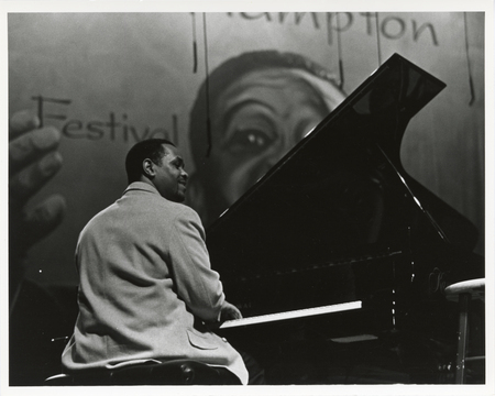 Black and white photograph of a man sitting at a piano. In the background is a large banner with a drawing of a man, partial text reading "Hampton Festival"