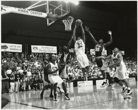 Black and white photograph of a men's basketball game in progress. Their uniforms read "IDAHO VANDALS" and "IONA". In the background, people sit or stand on stands.