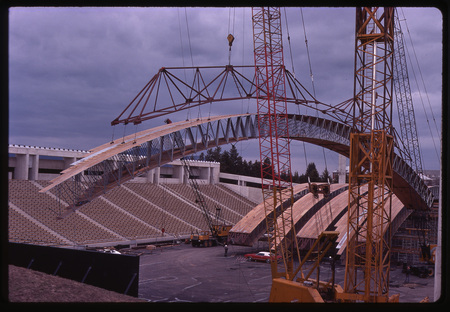 Color photograph of large wooden arches sitting on a stadium field and being maneuvered by large orange and red construction cranes.