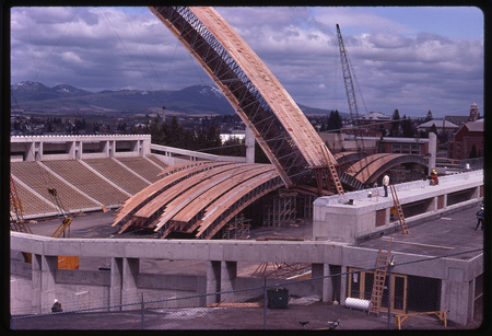 Color photograph of large wooden arches sitting on a stadium field and being maneuvered by large orange and red construction cranes.