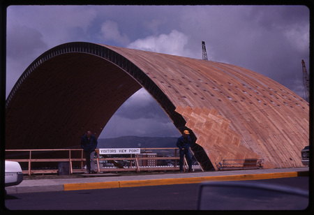 Color photograph of two men walking near a fence with a sign reading "Visitors View Point." In the background is a large domed building under construction.
