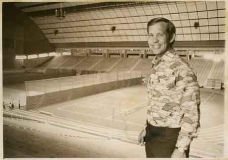 Black and white photograph of a man standing on an upper level of stadium stands, the enclosed stadium in the background.