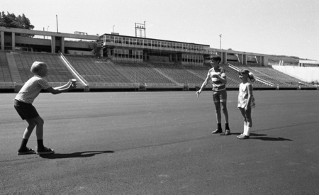 Black and white photograph of three children throwing a can to one another on the field of an open air stadium.