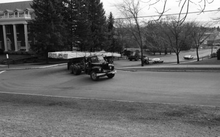 Black and white photograph of a truck driving through city streets, loaded with long, thin cargo.