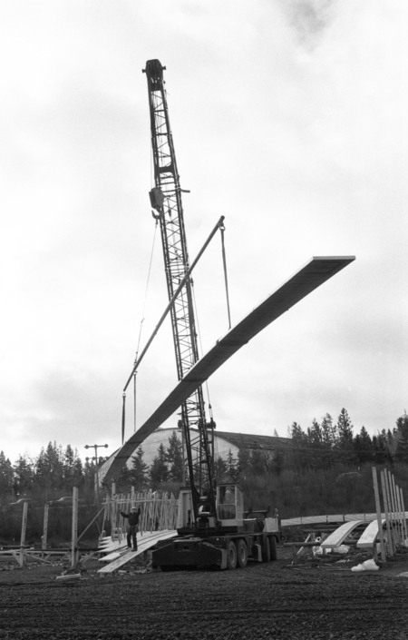 Black and white photograph of a construction crane maneuvering a wooden beam, directed by a worker on the ground.