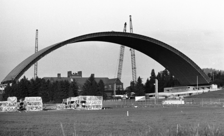 Black and white photograph of an arched wooden roof under construction. In the foreground are construction materials stacked on the ground. In the background is another building.
